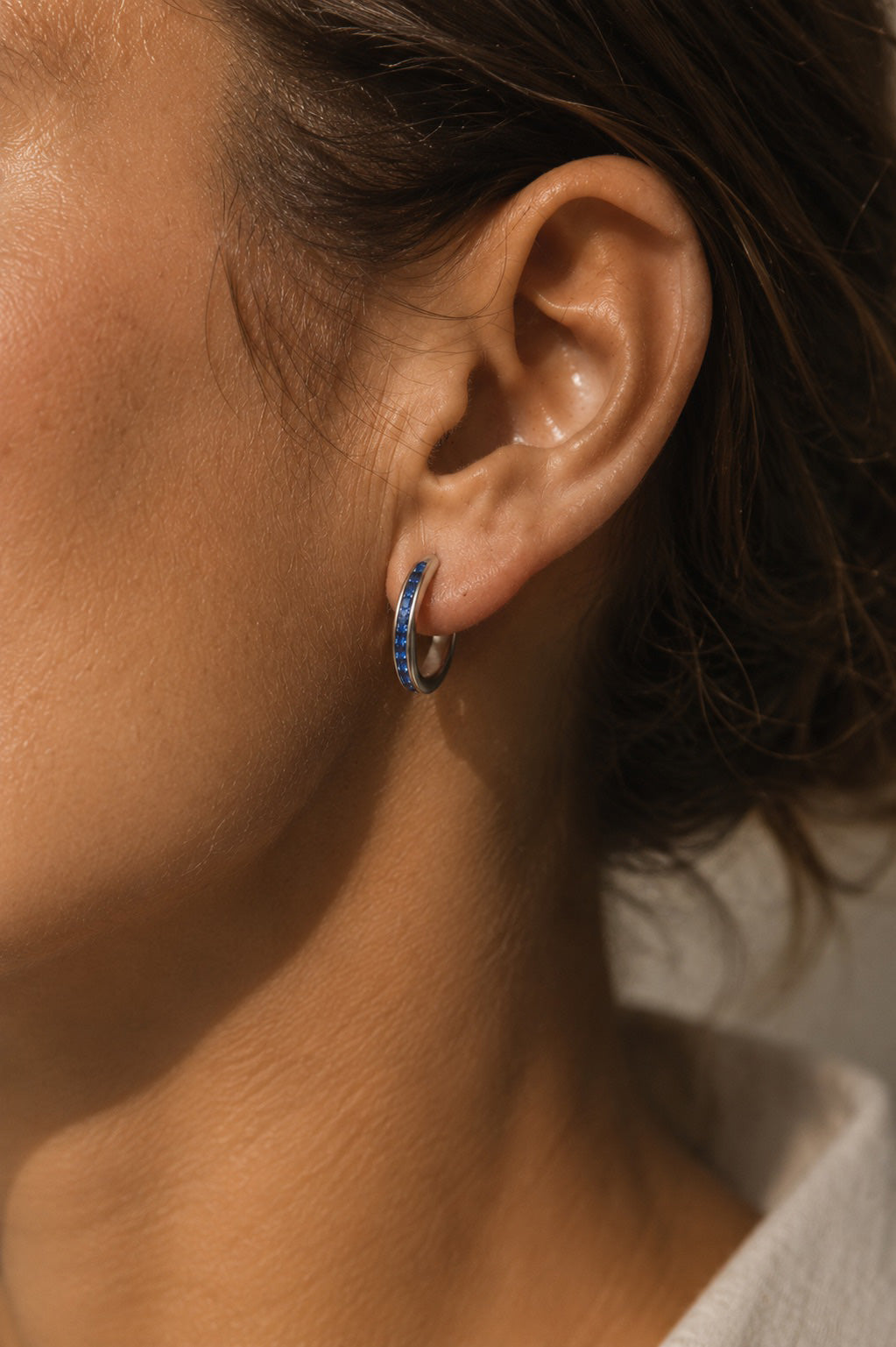 Close-up of an ear with a sterling silver hoop earring with natural sapphire beads  against a neutral background.
