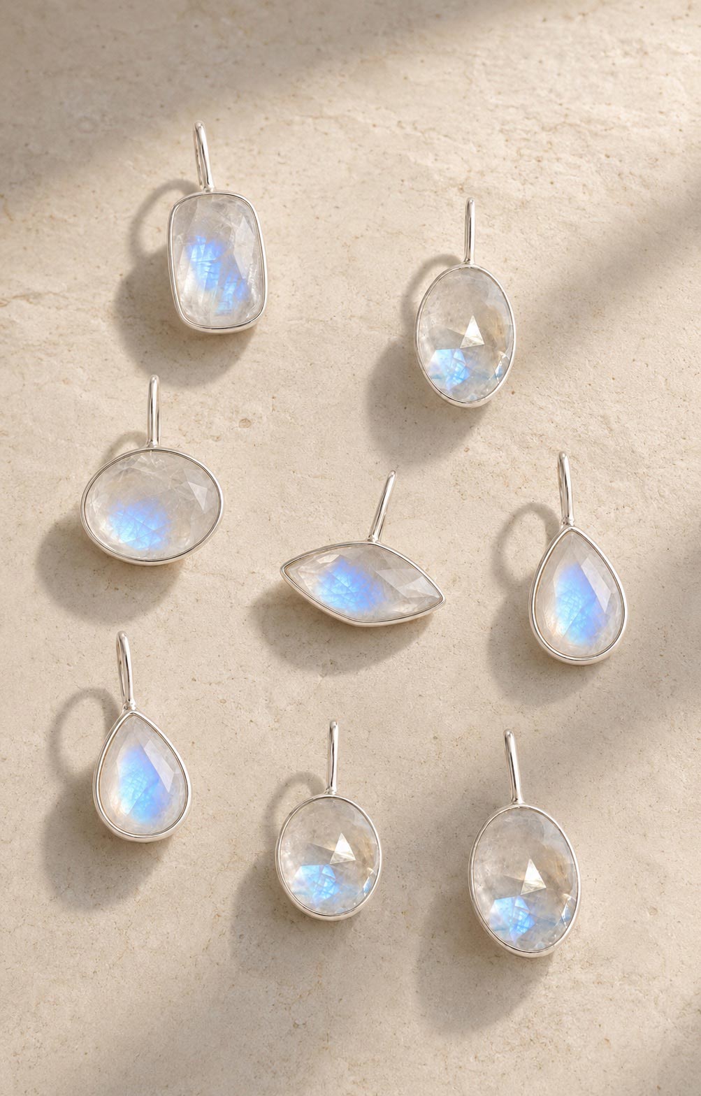 Seven sterling silver pendants with iridescent moonstones in teardrop, oval, and marquise shapes, arranged on a textured beige surface with soft shadows by Rya Atelier. 
